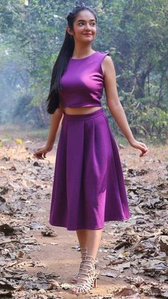 a woman in a purple dress is standing on a dirt path with trees and leaves behind her