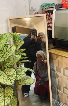 two women taking a selfie in front of a mirror with a plant behind them