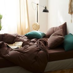 an unmade bed with brown and teal pillows on it in front of a window