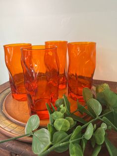 three orange glasses sitting on top of a wooden tray next to green leaves and greenery