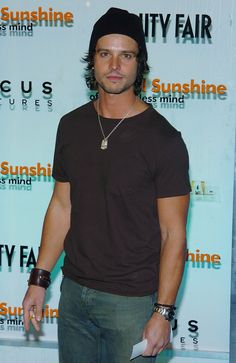 a man standing in front of a sign wearing a black hat and brown t - shirt