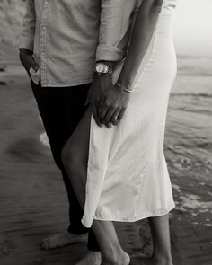 a man and woman holding hands while standing on the sand at the beach in black and white