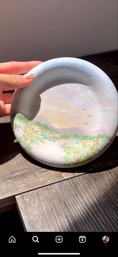 a hand holding a painted plate on top of a wooden table next to a window