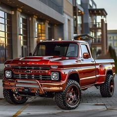 a red pickup truck parked in front of a building with large tires on it's rims