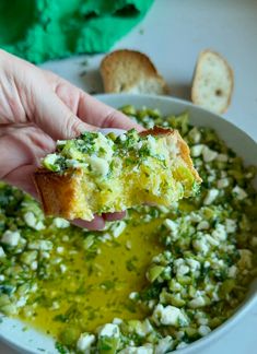 a hand holding a piece of bread over a bowl of green pesto and feta cheese