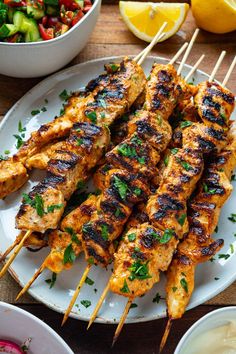 grilled chicken skewers on a white plate with lemons and salad in the background