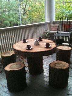 a wooden table and four stools on a porch with wood logs in the foreground