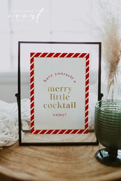 a merry little cocktail sign sitting on top of a wooden table next to a vase