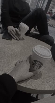 two people sitting at a table with coffee cups in their hands and one holding a cell phone