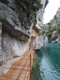 a wooden walkway leading to a body of water next to a rocky cliff face with trees growing on it