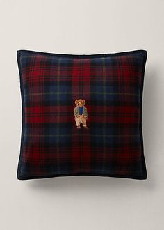 a red and blue plaid pillow with a teddy bear embroidered on the front, sitting against a white wall