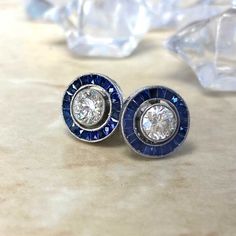 This pair of vintage style stud earrings features round brilliant cut diamonds with a sapphire halo. The total approximate diamond weight is 1.00 carats with J color and VS clarity. These earrings are platinum, with push-backs. ✦ DIAMOND SPECIFICATIONS: Diamond Cut: Brilliant Cut Diamond Weight 1.00 Carats Total ✦ ENGAGEMENT RING SPECIFICATIONS: Ring Material: Platinum Stones: Diamond ✦ WHAT COMES IN YOUR SHIPMENT: - Your Engagement Ring - Quality Ring Box - Jewelry Cleaner - UGL Certificate ✦ W Luxury Sapphire Diamond Earrings With Halo Design, Classic Sapphire-colored Cubic Zirconia Diamond Earrings, Vintage Round Platinum Diamond Earrings, Vintage Platinum Round Diamond Earrings, Sapphire Earrings With Diamond Accents, Classic Sapphire Earrings For Anniversary, Gia Certified Blue Diamond Round Earrings, Gia Certified Blue Diamond Earrings, Classic Sapphire Earrings With Halo Setting