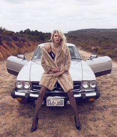 a woman sitting on top of a car in the dirt with her legs crossed and wearing high heels