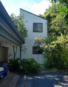 a white building with trees and bushes around it's corner in front of a blue car