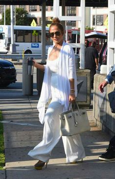 a woman is walking down the street carrying a purse and wearing white pants with yellow heels