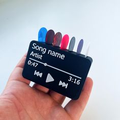 a hand holding a small black sign with colored markers on it that says song name artist