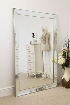 a white dresser sitting next to a mirror on top of a carpeted floor in front of a vase with flowers