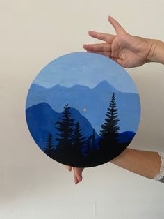 a person holding up a painted plate with mountains and trees in the background on it