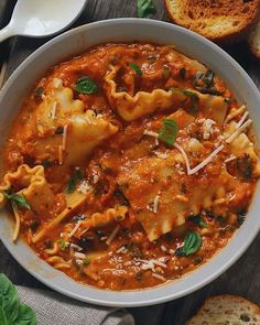 a white bowl filled with ravioli and sauce next to slices of bread on a wooden table