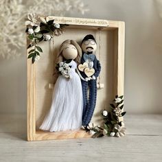 a wooden frame with two dolls in it and some flowers on the table next to it