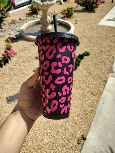 a person holding up a pink and black cup in front of a fountain with flowers