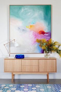 a painting hanging on the wall next to a wooden cabinet with vases and flowers