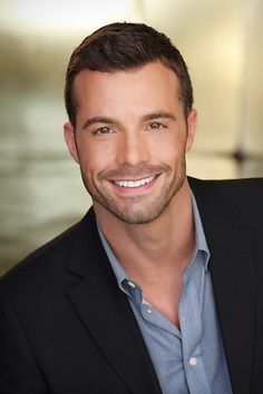 a smiling man in a black jacket and blue shirt is looking at the camera with a smile on his face