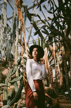 a woman standing in front of cactus trees wearing a hat and long sleeved top