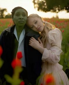 two people are standing in a field with flowers and trees behind them, one is hugging the other