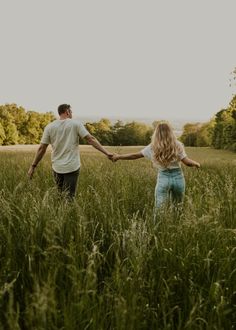 a man and woman holding hands while walking through tall grass in a field at sunset