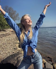 a woman standing on top of a rocky beach next to the ocean with her arms in the air