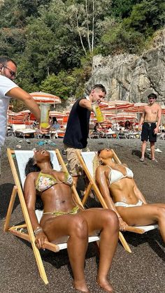 two women in bikinis are lounging on the beach while one man stands next to them