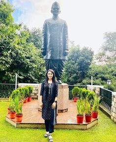 a woman standing in front of a statue with potted plants and trees behind her