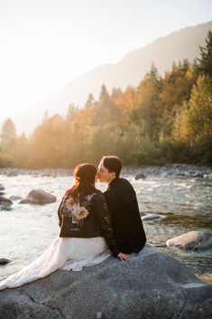 a man and woman sitting on top of a rock next to a river