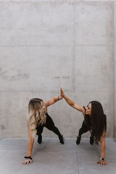 two women are doing handstands in front of a concrete wall and one is holding the other's hand