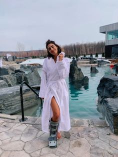 a woman is standing in front of a pool wearing boots and a white shirt dress