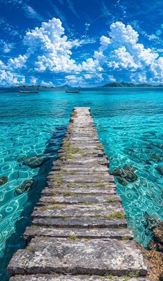 a long wooden dock in the middle of clear blue water with clouds above and an island in the distance