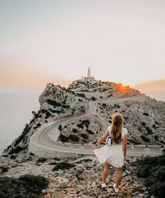 a woman standing on the side of a road next to a cliff with a lighthouse in the background