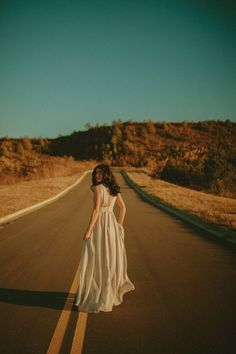 a woman in a white dress is standing on the side of an empty road with her hands behind her back