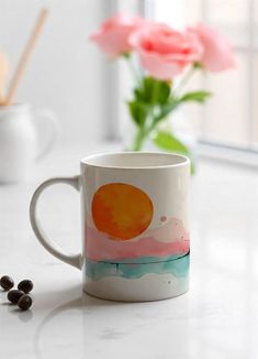 a coffee mug sitting on top of a white counter next to some black beans and pink flowers