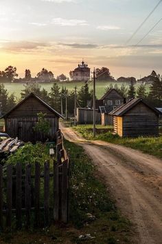 a dirt road runs between two small wooden buildings in the distance, with a field and trees behind it