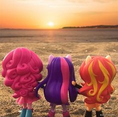 three little toy dolls are standing in the sand near the ocean at sunset or sunrise