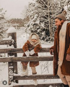 two people standing next to each other in the snow near a fence with trees behind them