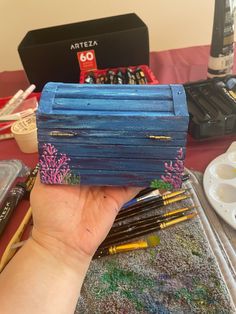 a hand holding up a blue box that has been painted with paint and pencils on it