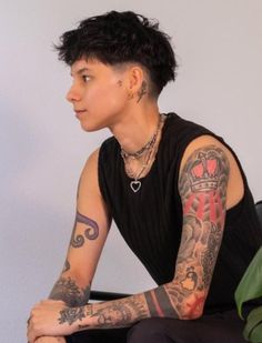 a man with tattoos sitting on the ground next to a plant and wearing a black shirt