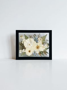 a white flower arrangement in a black frame on top of a table next to a wall