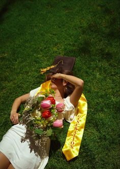 a woman laying in the grass with flowers and a graduation sash around her neck, covering her face