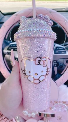 a person holding up a pink hello kitty cup in their hand and steering wheel behind her