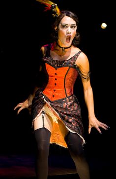 a woman in an orange and black dress with her hands out to the side while standing on a stage