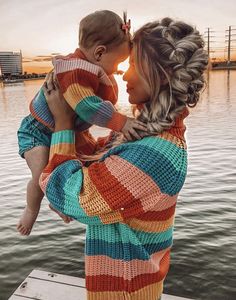 a woman holding a baby in her arms while standing on a dock near the water